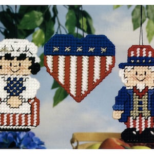 May include: Three handmade felt ornaments with a patriotic theme. A heart-shaped ornament with red and white stripes and blue stars, a woman wearing a white dress and a red, white, and blue hat, and a man wearing a blue jacket and a red, white, and blue hat.