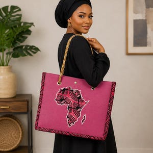 May include: A magenta tote bag with a gold rope handle and a decorative African continent design. The bag has a patterned border and a button closure. The bag is carried over the shoulder.