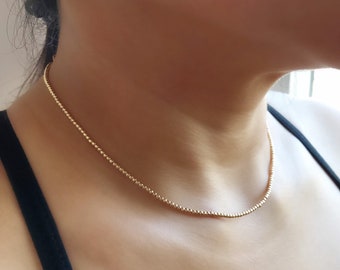 May include: A delicate gold chain necklace with a beaded design. The necklace is worn by a person with a light complexion and dark hair.