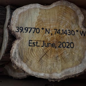 May include: Wooden tree slice with the coordinates 39.9770° N, 74.1430° W and the text "Est. June, 2020" engraved in black. The wood has a light brown color with visible grain and bark.