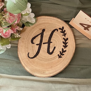 May include: A wooden table number holder with the number 4, and a wooden round place card with the letter "H" and a leaf design. A bouquet of pink and white flowers is in the background.