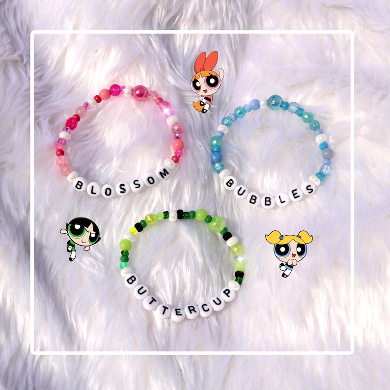 Op de afbeelding: Drie kleurrijke kralenarmbanden met de namen "Blossom", "Bubbles" en "Buttercup" gespeld in witte letters. Elke armband heeft een cartoonfiguur uit de Powerpuff Girls.