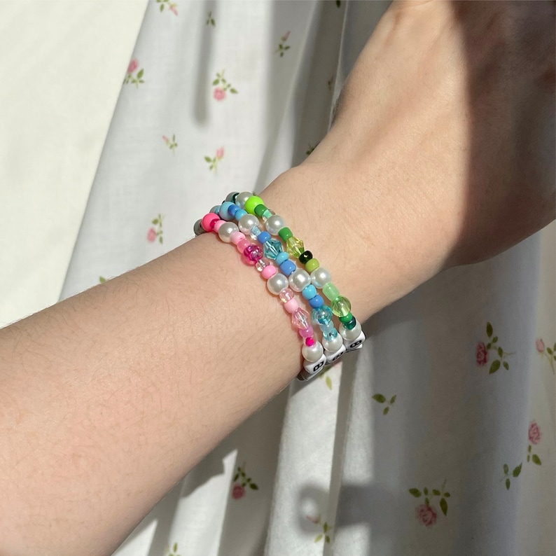 Op de afbeelding: Een stapel van drie kralenarmbanden om een pols. De armbanden zijn gemaakt van witte, blauwe, roze en groene kralen. De kralen zijn geregen op elastiek.