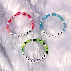 Op de afbeelding: Drie kralenarmbanden met witte letterkralen die de namen "Blossom", "Bubbles" en "Buttercup" spellen. De armbanden zijn gemaakt van roze, blauwe en groene kralen.