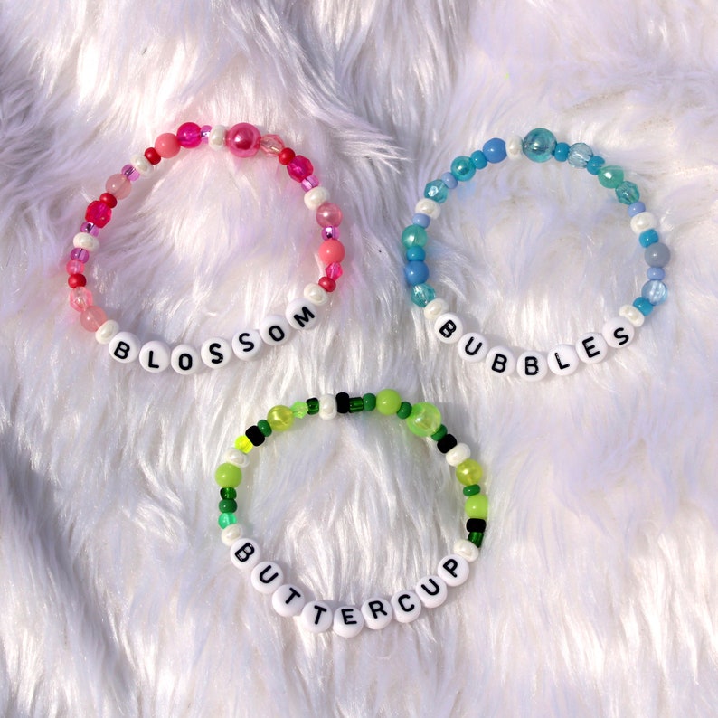 Op de afbeelding: Drie kralenarmbanden met witte letterkralen die de namen "Blossom", "Bubbles" en "Buttercup" spellen. De armbanden zijn gemaakt van roze, blauwe en groene kralen.