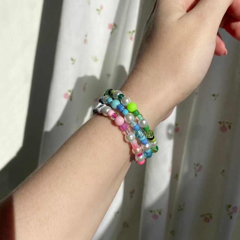 Op de afbeelding: Drie kleurrijke kralenarmbanden met witte parels. De armbanden zijn gestapeld op de pols van een persoon.