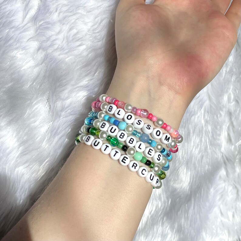 Op de afbeelding: Een stapel van zes kleurrijke kralenarmbanden met de woorden "Blossom", "Bubbles" en "Buttercup" gespeld in witte letters.