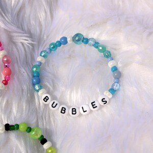 Op de afbeelding: Een blauwe en witte kralen armband met het woord "BUBBLES" gespeld in witte kralen.