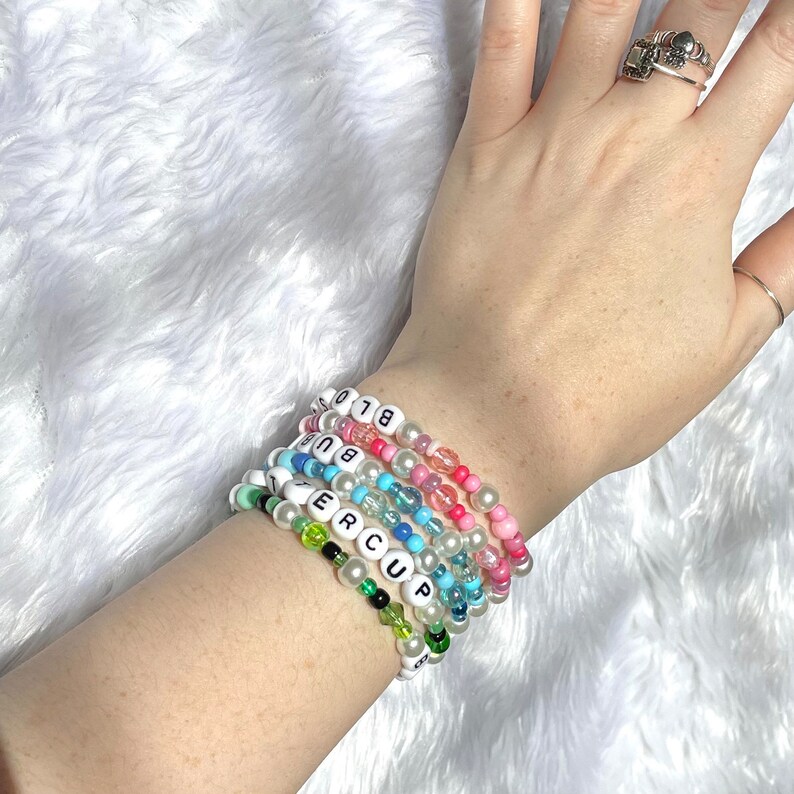 Op de afbeelding: Een stapel kleurrijke kralenarmbanden met witte letterkralen die "BIG" en "SUPER CUP" spellen.