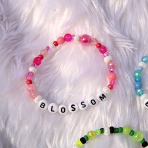 Op de afbeelding: Een roze en witte kralen armband met het woord "BLOSSOM" gespeld in witte letters.