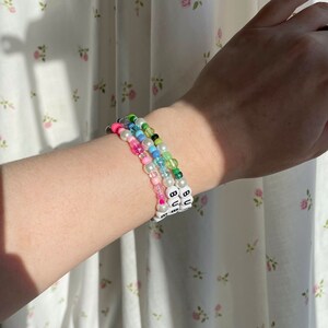 Op de afbeelding: Drie kralenarmbanden met witte kralen en zwarte letters die "BU" op elke armband spellen. De armbanden zijn op elkaar gelegd op de pols van een persoon en hebben verschillende gekleurde kralen, waaronder roze, blauw, groen en wit.