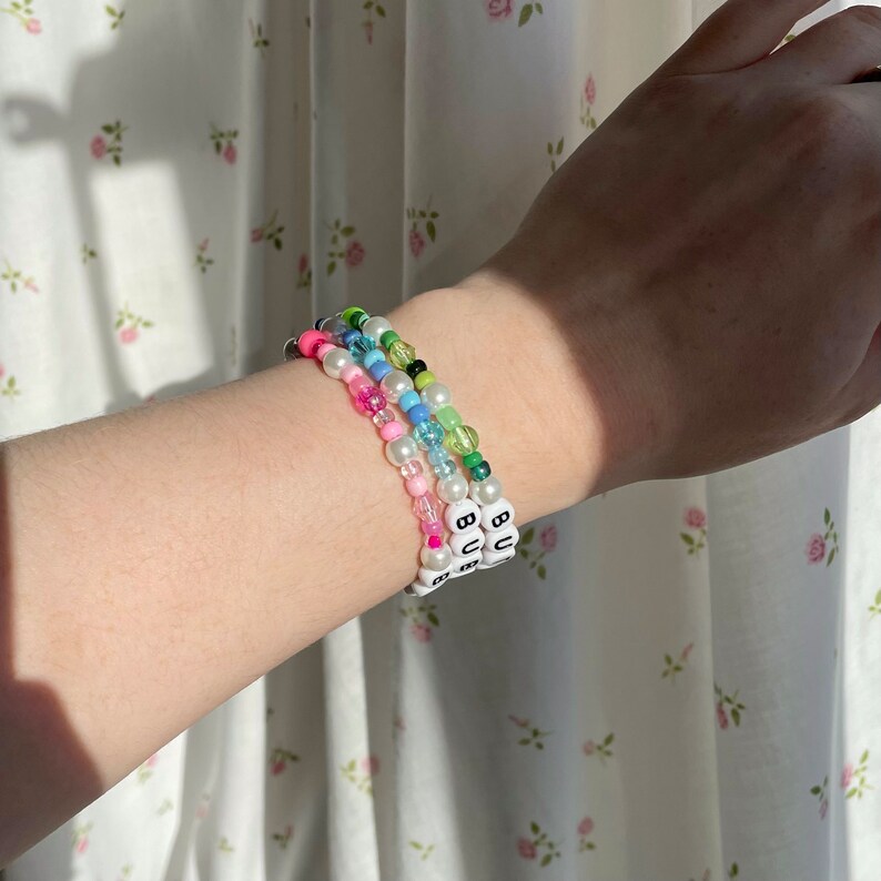 Op de afbeelding: Drie kralenarmbanden met witte kralen en zwarte letters die "BU" op elke armband spellen. De armbanden zijn op elkaar gelegd op de pols van een persoon en hebben verschillende gekleurde kralen, waaronder roze, blauw, groen en wit.