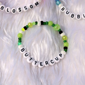 Op de afbeelding: Een groene, gele en witte kralen armband met het woord "BUTTERCUP" gespeld in witte kralen.