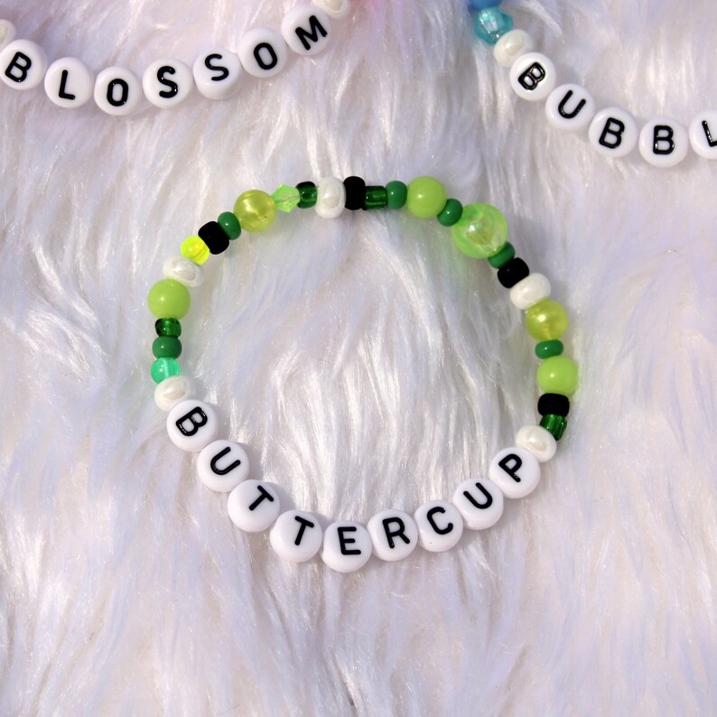 Op de afbeelding: Een groene, gele en witte kralen armband met het woord "BUTTERCUP" gespeld in witte kralen.