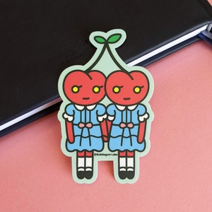 May include: Two red cherries with faces and blue dresses holding hands. The cherries have green stems and leaves. The text "Bubblegum Labs" is at the bottom of the sticker.