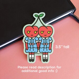 May include: Two red cherries with faces and blue dresses holding hands. The cherries are 3.5 inches tall.  The sticker is on a pink surface.  Please read description for additional good info :)