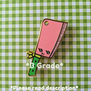 May include: Pink and green enamel pin shaped like a cartoon cleaver with a gold outline and glitter accents. The pin has a small yellow bow and says "*B Grade*" and "*Please read description*" in black text.