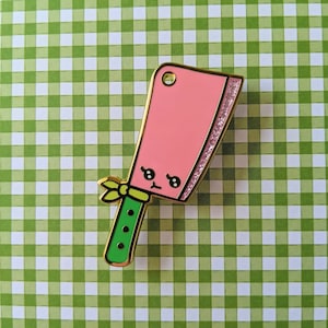 May include: A pink cleaver enamel pin with a gold outline and a glittery edge. The cleaver has a cute face and a green handle with gold accents. The pin is set against a green and white checkered background.