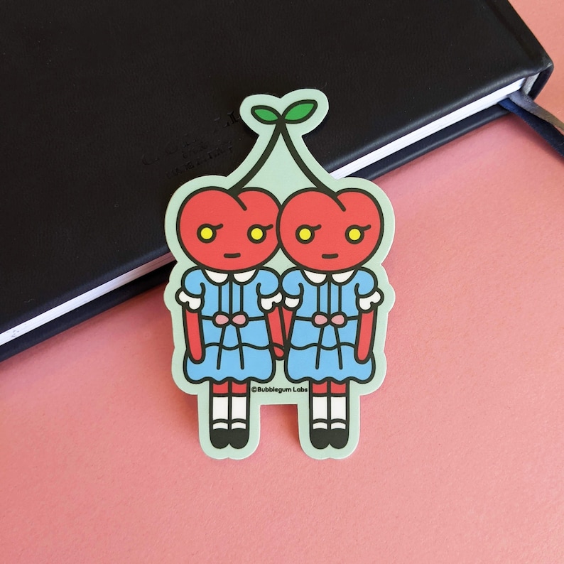 May include: Two red cherries with faces and blue dresses holding hands. The cherries have green stems and leaves. The text "Bubblegum Labs" is at the bottom of the sticker.