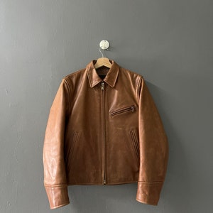 Puede incluir: Una chaqueta de cuero marrón con cierre de cremallera y dos bolsillos en la parte delantera. La chaqueta está colgada de un gancho contra una pared gris.