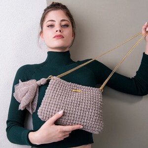 以下が含まれることがあります： 濃い緑色のタートルネックセーターを着た女性が、金色のチェーンストラップが付いた薄い茶色のクロシェ編みのハンドバッグを持っています。バッグには、前面に小さな金色のアクセントがあります。