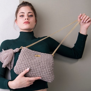 以下が含まれることがあります： 緑色のタートルネックセーターを着た女性が、金色のチェーンストラップが付いた薄い茶色のクロシェ編みのハンドバッグを持っています。バッグには、「Lilla」と書かれた小さな金色のタグが付いています。