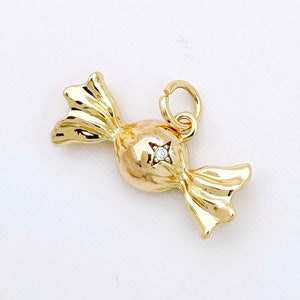 Puede incluir: Un amuleto de caramelo de color dorado con un solo diamante transparente en el centro. El amuleto tiene forma de caramelo envuelto con dos extremos puntiagudos.