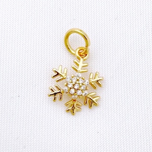 Op de afbeelding: Een gouden sneeuwvlok-bedel met heldere kristallen. De bedel heeft een lus aan de bovenkant om aan een ketting of armband te bevestigen.