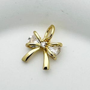 May include: A gold-coloured bow pendant with two clear, sparkling gemstones. The bow design features curved lines and a loop for attaching to a chain. The pendant is set against a white background.