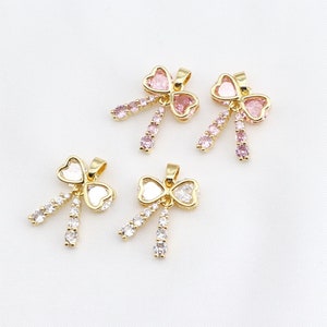 May include: Three gold bow-shaped pendants with clear and pink crystal embellishments. The pendants are on a white background.