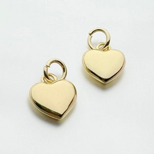 Op de afbeelding: Twee gouden hartvormige bedels met een lus aan de bovenkant om aan een ketting of armband te bevestigen.
