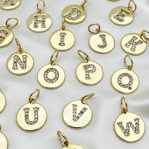 以下が含まれることがあります： アルファベットの異なる文字が刻まれた、金色のイニシャルチャームのコレクション。文字は小さな透明なクリスタルで装飾されています。チャームは丸く、チェーンやブレスレットに取り付けるための小さなループが上部に付いています。