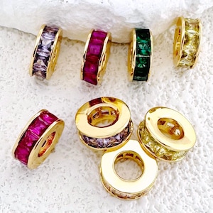 Könnte beinhalten: Eine Sammlung goldfarbener, kreisförmiger Schmuckstücke mit farbigen Edelsteinen. Die Edelsteine sind in den Farbtönen Rot, Grün, Lila und Gelb gehalten. Der Schmuck wird auf einer weißen, strukturierten Oberfläche präsentiert.