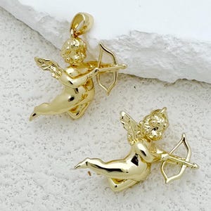 May include: Two gold-colored cupid pendants, each holding a bow and arrow. The pendants feature detailed wings and flowing hair, with a loop at the top for a chain. The background is a textured white surface.