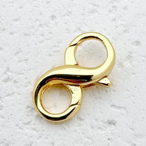 May include: A gold-colored lobster clasp, a jewelry component, is shown against a textured white background. The clasp has a curved design with two circular openings and a lever for secure closure. The metal has a shiny finish.