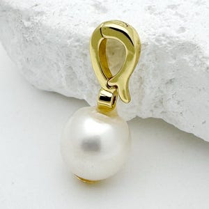 Puede incluir: Un colgante dorado con una gran perla blanca. El colgante tiene un diseño curvo en forma de gancho en la parte superior, y la perla es redonda y tiene una superficie lisa. La joya está colocada sobre un fondo blanco texturizado.