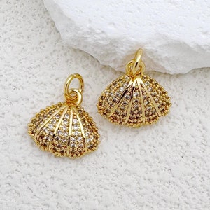 Puede incluir: Dos colgantes dorados con forma de concha marina con cristales transparentes y marrones. Los colgantes están sobre una superficie blanca texturizada. Cada colgante tiene un pequeño lazo en la parte superior para sujetarlo. El diseño de concha es detallado y elegante.