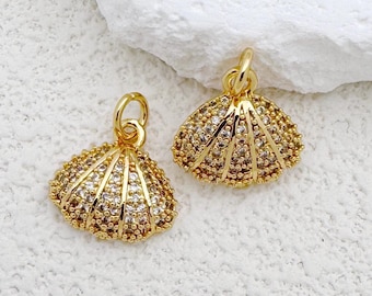 May include: Two gold-coloured seashell charms with clear and brown crystals. The charms are on a white textured surface. Each charm has a small loop at the top for attaching to a necklace or bracelet. The seashell design is detailed and elegant.
