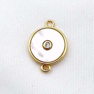 Könnte beinhalten: Goldfarbener Anhänger mit einem weißen Perlmuttkreis und einem kleinen klaren Strassstein in der Mitte. Der Anhänger hat zwei Ösen auf der Rückseite zum Anbringen an einer Halskette oder einem Armband.
