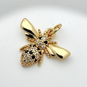 Pode incluir: Um pingente de abelha dourada com detalhes em cristais pretos e transparentes. A abelha tem asas detalhadas e um pequeno laço para prender a um colar ou pulseira. O pingente está sobre um fundo branco.