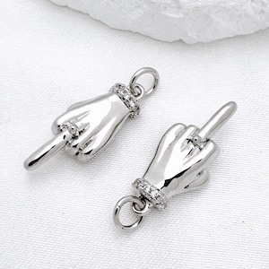 Peut inclure: Deux pendentifs argentés en forme de mains faisant un doigt d'honneur. Chaque pendentif est orné d'une petite boucle en haut et d'une bande de petits strass transparents autour du poignet. Sur fond blanc.