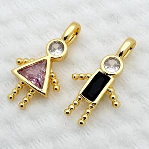 May include: Two gold-colored pendant charms in the shape of stick figures. One has a pink triangle body and a clear gem head, the other has a black rectangle body and a clear gem head. Both have small gold bead arms and legs.
