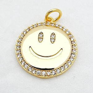 Op de afbeelding: Goudkleurige hanger met een lachend gezicht. De ronde hanger heeft een rand van kleine, heldere kristallen. Het smiley-gezicht heeft twee kleine, ovale ogen en een gebogen mond. Bovenop zit een kleine lus voor een ketting.