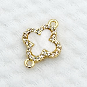 May include: A gold-colored clover charm with a white center and a border of small, clear crystals. The charm has two small loops for attaching to jewelry. The clover is a four-leaf design.