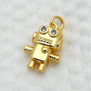 May include: A gold robot charm with a loop for hanging. The robot has two round, clear gems for eyes and a heart-shaped detail on its chest. The charm is small and detailed, with a polished finish.