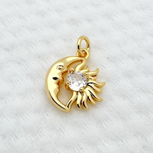 May include: A gold-colored pendant featuring a crescent moon embracing a sun with a central clear gemstone. The pendant has a small loop for attaching to a chain. The design is detailed and has a polished finish.