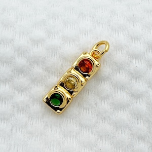Puede incluir: Un dije dorado con un diseño de semáforo. El dije tiene tres piedras preciosas de colores: rojo, amarillo y verde. El dije es rectangular con esquinas redondeadas y un pequeño lazo en la parte superior.