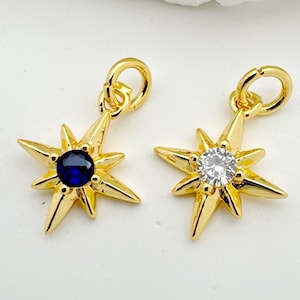 Könnte beinhalten: Zwei goldfarbene Stern-Charms. Ein Charm hat einen dunkelblauen Edelstein, der andere einen klaren, funkelnden Edelstein. Jeder Charm hat oben eine kleine Schlaufe zum Befestigen an Schmuck.