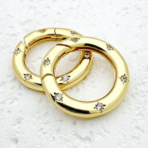 Può includere: Due chiusure rotonde color oro con apertura a cerniera. Ogni chiusura è adornata da piccoli abbellimenti scintillanti a forma di stella. Le chiusure sono poste su uno sfondo bianco strutturato, creando un contrasto che ne evidenzia il design.