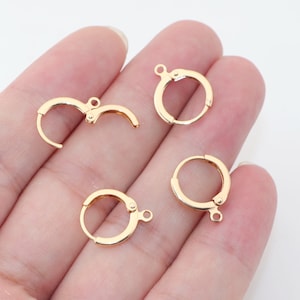 Könnte beinhalten: Vier goldfarbene Ohrringverschlüsse mit Hebelverschluss. Die Verschlüsse sind klein, kreisförmig und haben ein Scharnierdesign für eine einfache Befestigung. Jeder Verschluss hat eine kleine Schlaufe zum Anbringen eines Anhängers oder einer Perle. Weißer Hintergrund.